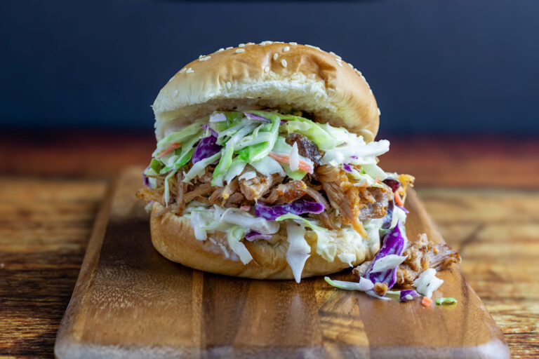 A close-up shot of Southern coleslaw piled high on a pulled pork sandwich, showing the creamy dressing glistening on crisp shredded cabbage with the soft bun visible beneath, set on a rustic wooden board