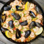 Overhead shot of a large Spanish paella pan filled with golden saffron rice, shrimp, mussels, chicken, and chorizo, garnished with lemon wedges and fresh parsley on a rustic wooden table