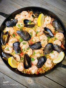 Overhead shot of a large Spanish paella pan filled with golden saffron rice, shrimp, mussels, chicken, and chorizo, garnished with lemon wedges and fresh parsley on a rustic wooden table