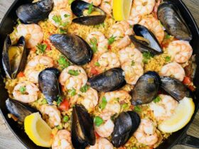 Overhead shot of a large Spanish paella pan filled with golden saffron rice, shrimp, mussels, chicken, and chorizo, garnished with lemon wedges and fresh parsley on a rustic wooden table