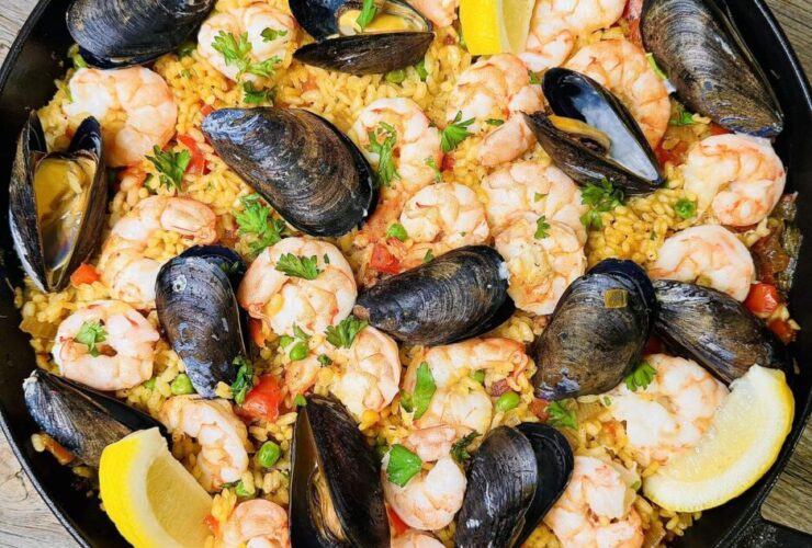 Overhead shot of a large Spanish paella pan filled with golden saffron rice, shrimp, mussels, chicken, and chorizo, garnished with lemon wedges and fresh parsley on a rustic wooden table