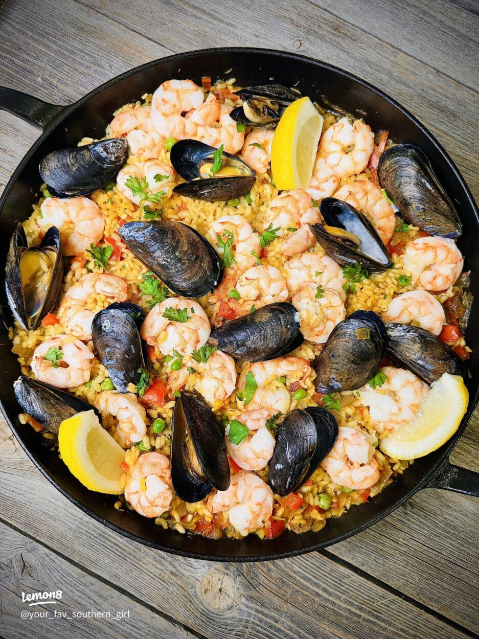 Overhead shot of a large Spanish paella pan filled with golden saffron rice, shrimp, mussels, chicken, and chorizo, garnished with lemon wedges and fresh parsley on a rustic wooden table