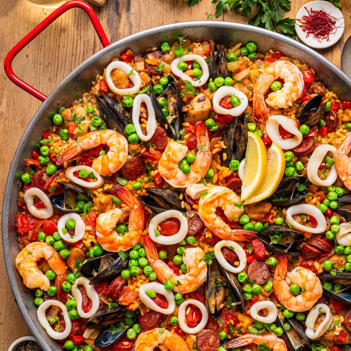 Flat lay of Spanish paella ingredients including saffron threads, Bomba rice, raw shrimp, mussels, chicken thighs, chorizo slices, smoked paprika, and fresh tomatoes on a marble surface