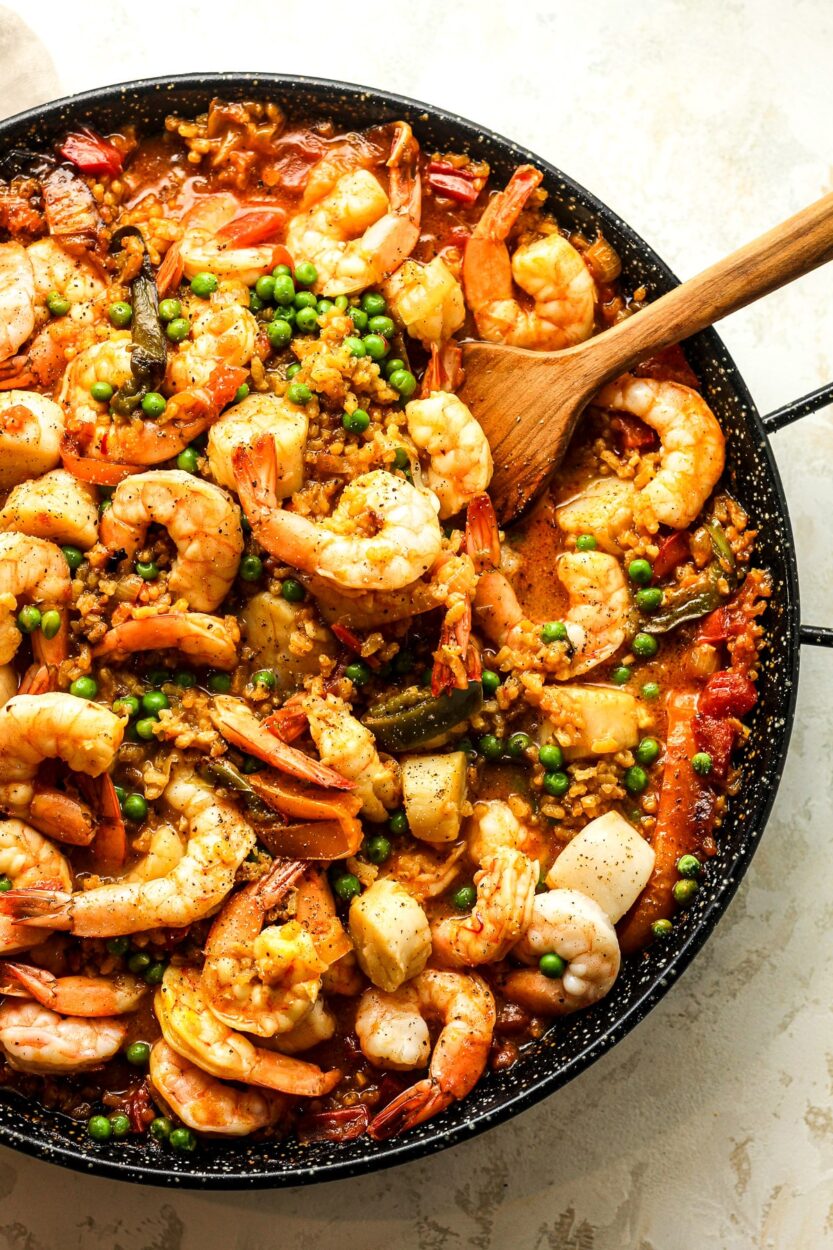 Close-up of a spoonful of Spanish paella being lifted from the pan, showing golden saffron rice, a plump shrimp, and a mussel, with the crispy socarrat visible on the bottom layer