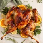 Beautifully plated whole spatchcock turkey on a large wooden serving board with fresh rosemary, thyme, sliced citrus, and pan drippings poured over the skin, golden brown and crispy
