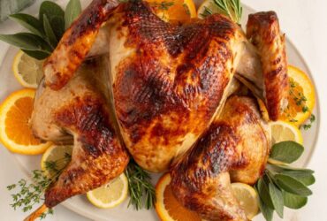 Beautifully plated whole spatchcock turkey on a large wooden serving board with fresh rosemary, thyme, sliced citrus, and pan drippings poured over the skin, golden brown and crispy