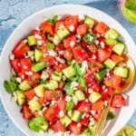Watermelon Feta Salad served on a large white platter with crumbled feta, fresh mint, cucumber slices, and a drizzle of honey and lime juice, photographed overhead on a rustic wooden table in bright summer light