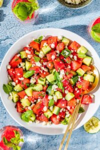 Watermelon Feta Salad served on a large white platter with crumbled feta, fresh mint, cucumber slices, and a drizzle of honey and lime juice, photographed overhead on a rustic wooden table in bright summer light