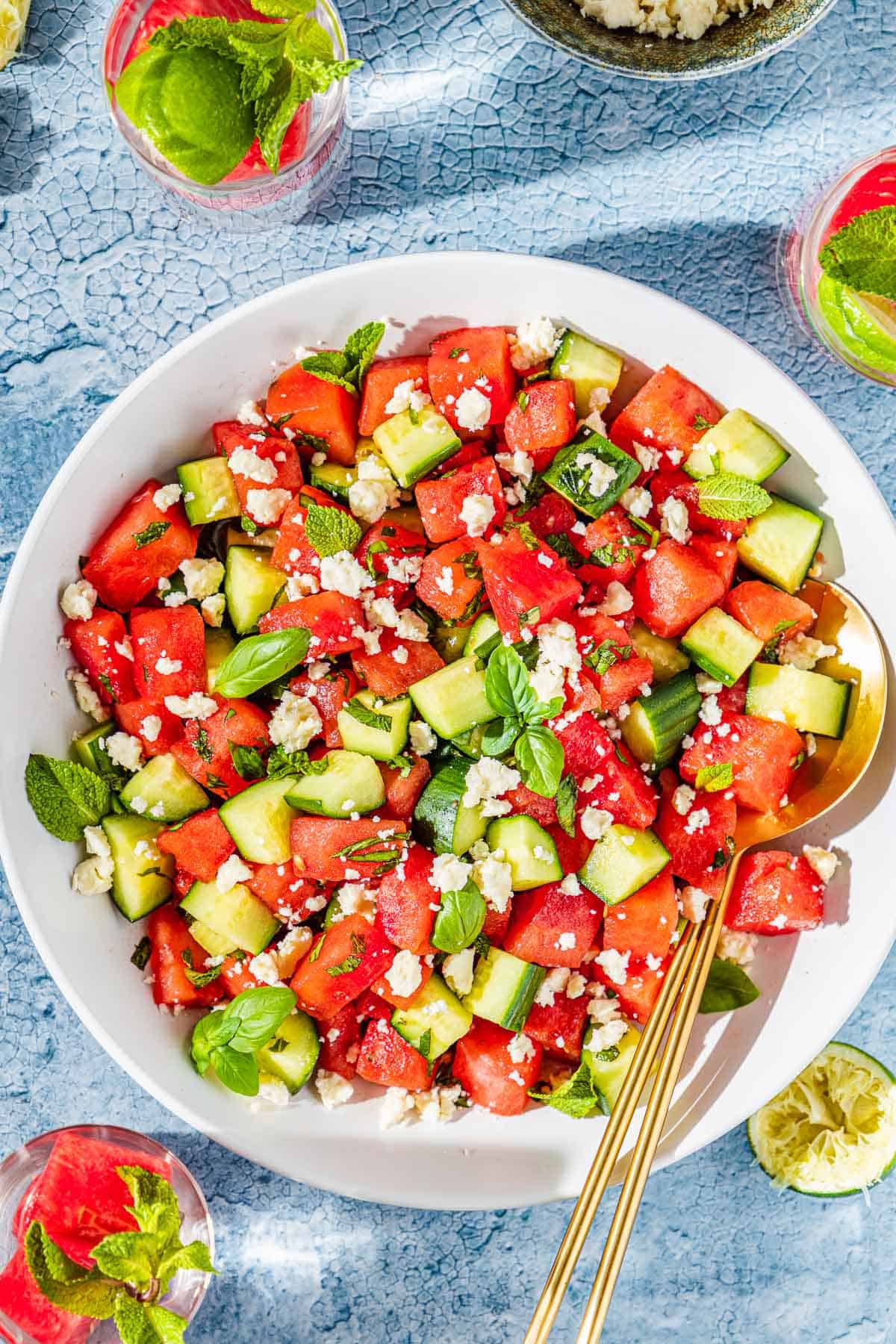 Watermelon Feta Salad served on a large white platter with crumbled feta, fresh mint, cucumber slices, and a drizzle of honey and lime juice, photographed overhead on a rustic wooden table in bright summer light