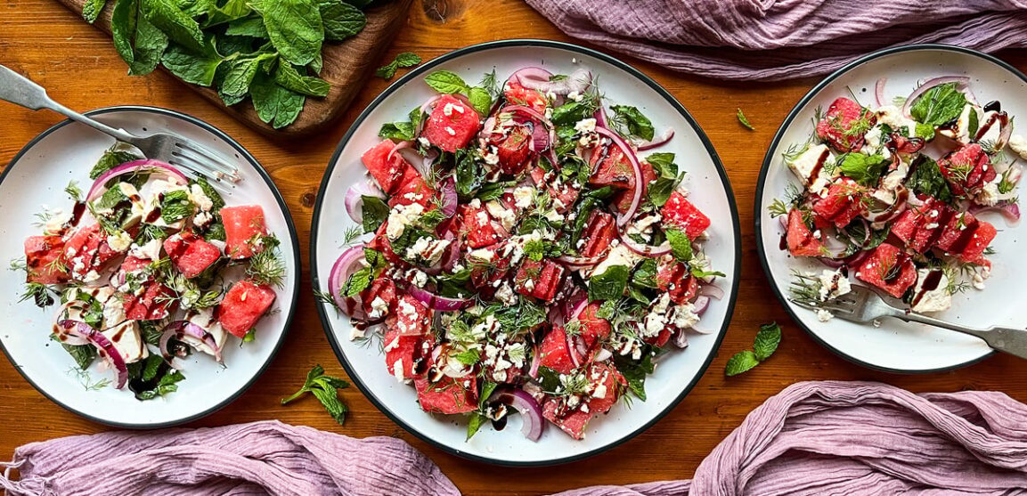 Close-up of a single serving of Watermelon Feta Salad in a shallow bowl, showing vibrant red watermelon chunks topped with crumbled white feta, torn mint leaves, and a glistening drizzle of honey and lime, with a fork resting beside it