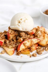Close-up of a single serving of apple crisp in a bowl, showing the tender cinnamon-spiced apple layers beneath the golden oat streusel, topped with a melting scoop of vanilla ice cream