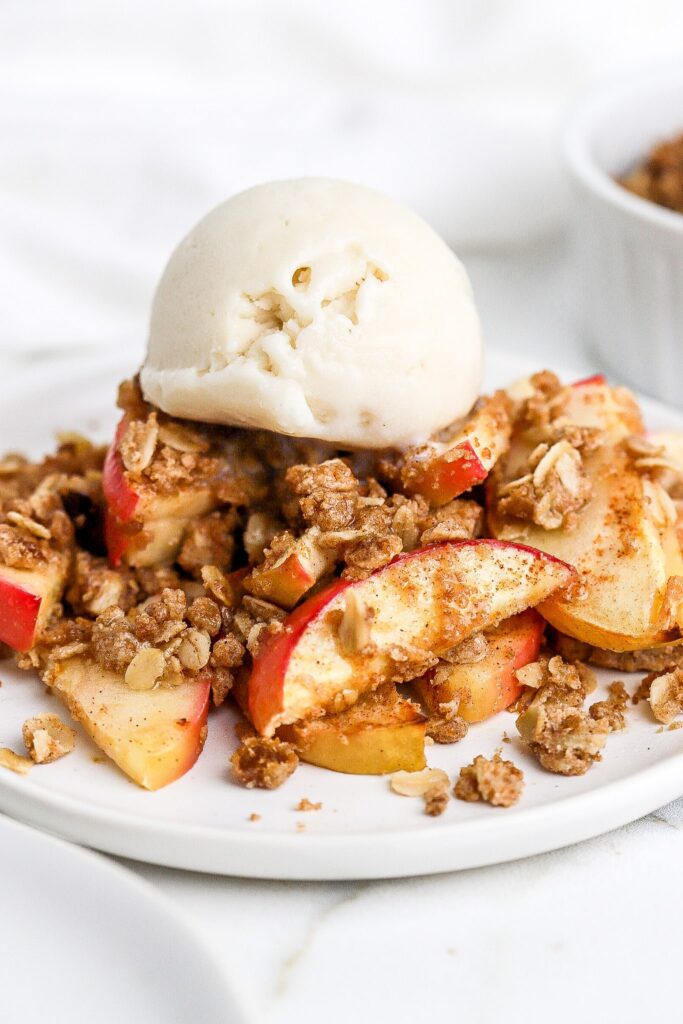 Close-up of a single serving of apple crisp in a bowl, showing the tender cinnamon-spiced apple layers beneath the golden oat streusel, topped with a melting scoop of vanilla ice cream