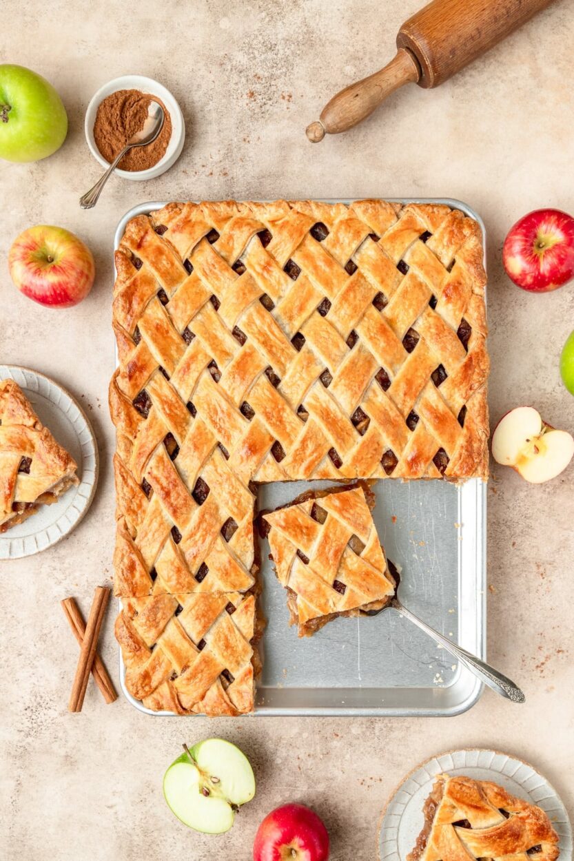 Golden apple slab pie on a sheet pan, sliced into squares with flaky top crust and cinnamon apple filling oozing at the edges, served on a rustic wooden board