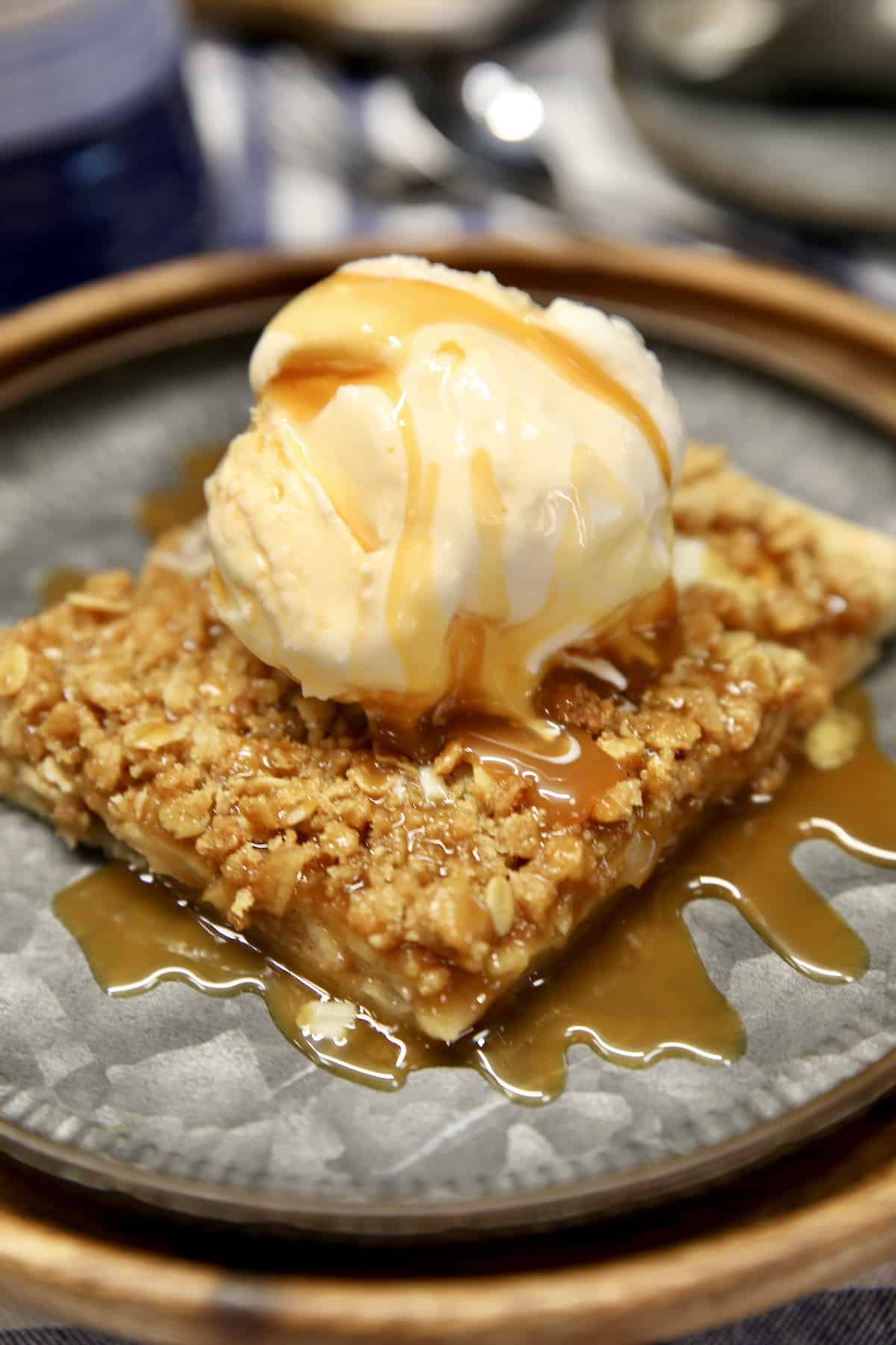 Close-up of a single apple slab pie square on a white plate, topped with a scoop of vanilla ice cream melting over the golden flaky crust, warm caramel apple filling visible at the cut edge