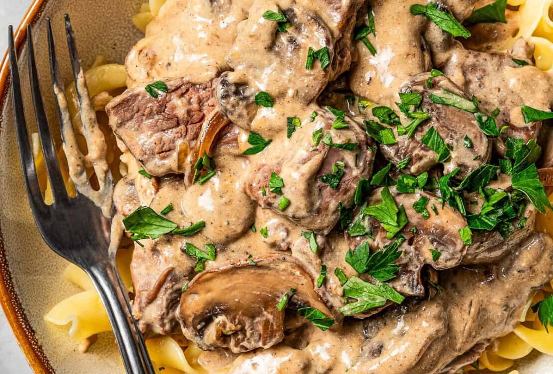 Beef stroganoff served over egg noodles in a wide bowl, topped with fresh parsley and cracked black pepper, rustic table setting