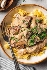 Beef stroganoff served over egg noodles in a wide bowl, topped with fresh parsley and cracked black pepper, rustic table setting