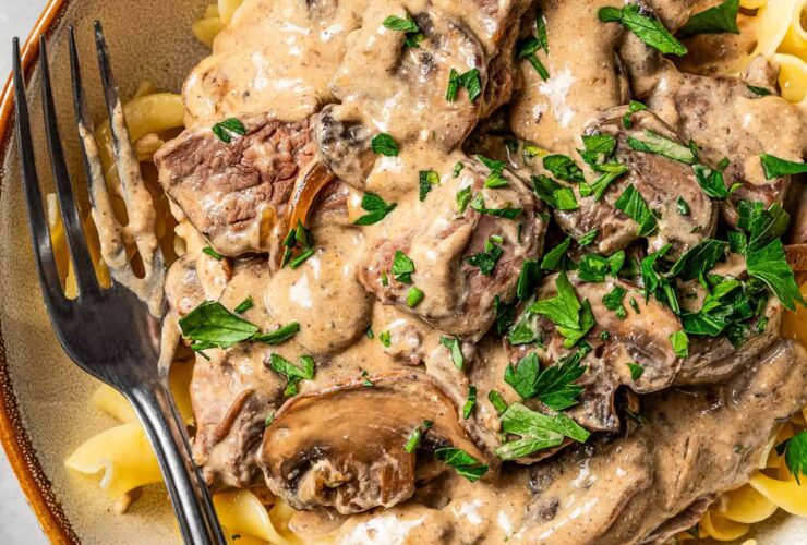Beef stroganoff served over egg noodles in a wide bowl, topped with fresh parsley and cracked black pepper, rustic table setting
