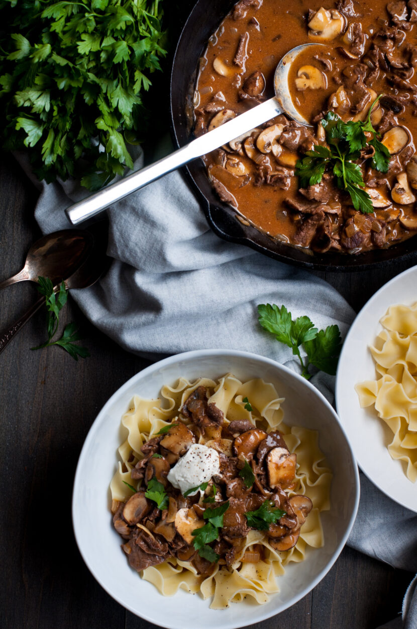 Sliced sirloin, cremini mushrooms, sour cream, beef broth, Dijon mustard, and egg noodles arranged on a wooden board — beef stroganoff ingredients flat lay