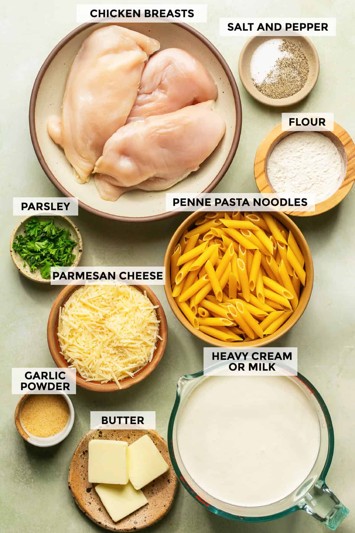 Ingredients for chicken alfredo bake laid out flat — penne pasta, shredded rotisserie chicken, heavy cream, garlic cloves, parmesan block, mozzarella, and butter on a marble surface