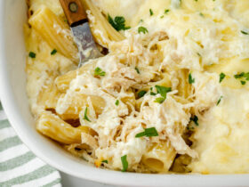 Close-up of a serving spoon lifting a cheesy, saucy portion of chicken alfredo bake from the casserole dish, showing the stretch of melted mozzarella and creamy parmesan sauce coating the penne pasta
