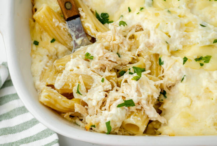 Close-up of a serving spoon lifting a cheesy, saucy portion of chicken alfredo bake from the casserole dish, showing the stretch of melted mozzarella and creamy parmesan sauce coating the penne pasta