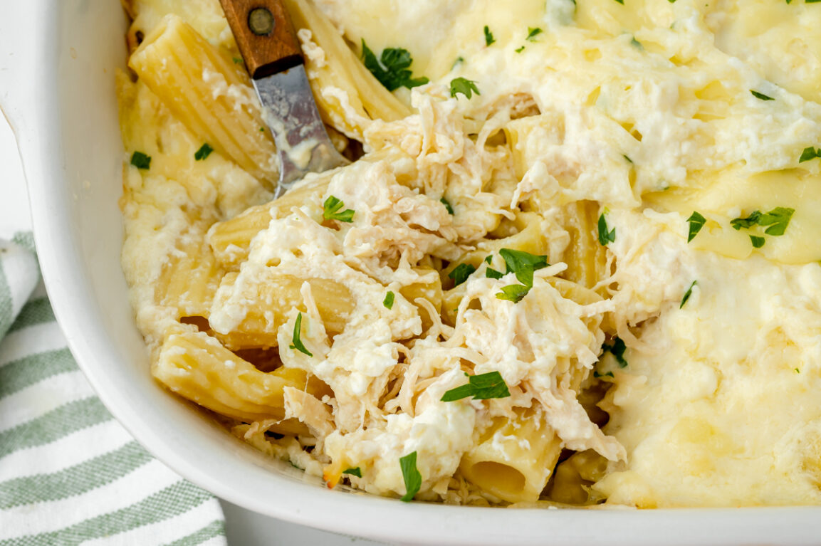 Close-up of a serving spoon lifting a cheesy, saucy portion of chicken alfredo bake from the casserole dish, showing the stretch of melted mozzarella and creamy parmesan sauce coating the penne pasta