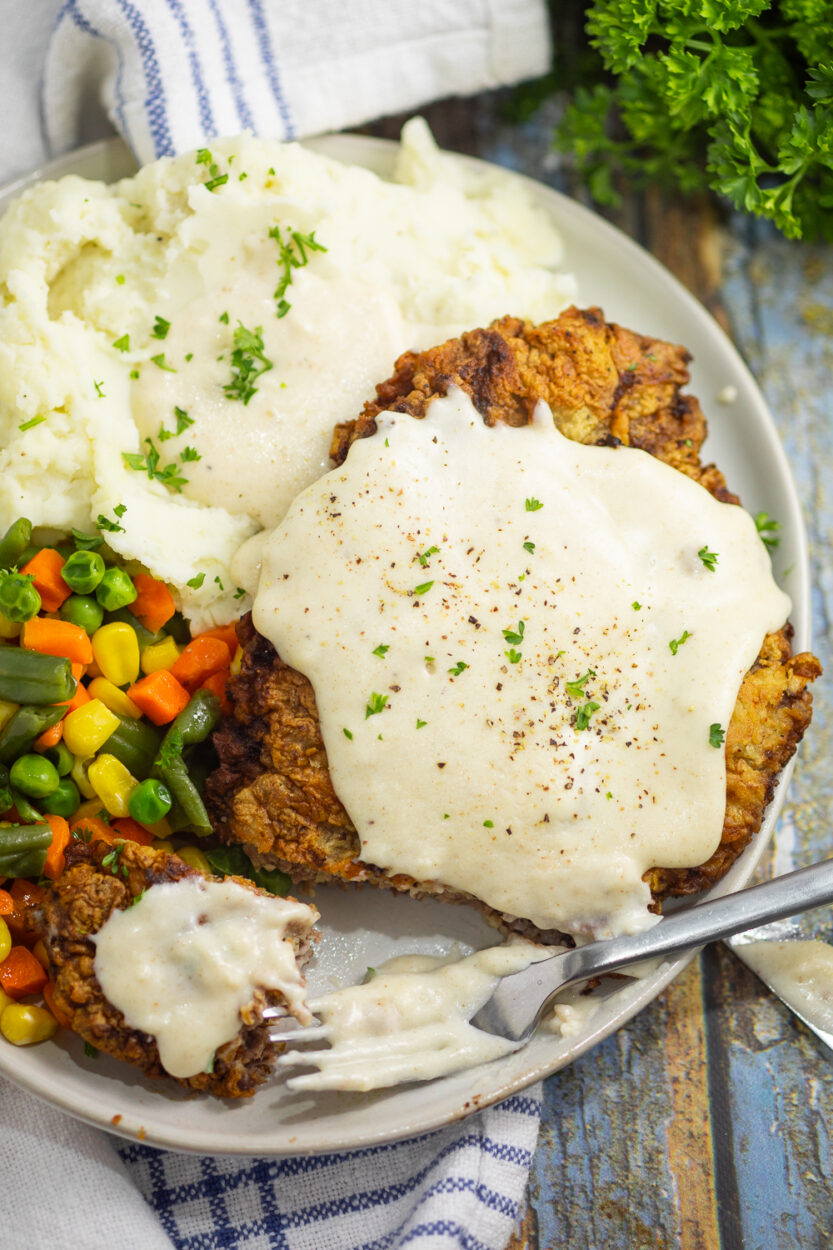 Chicken fried steak on a white plate smothered in peppery white gravy with mashed potatoes on the side