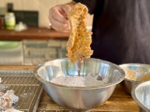 Cube steak being dredged through seasoned flour and egg wash on a wooden board with spices nearby