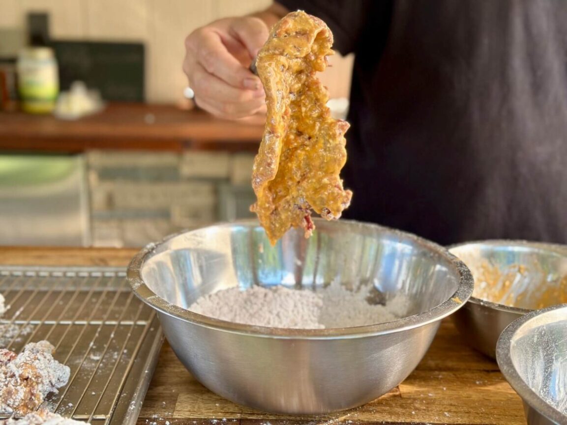 Cube steak being dredged through seasoned flour and egg wash on a wooden board with spices nearby