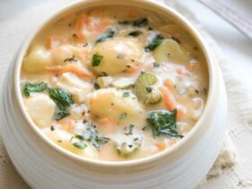 A bowl of creamy chicken gnocchi soup garnished with fresh thyme and a crack of black pepper, with soft potato gnocchi and shredded carrots visible in the rich cream broth, photographed on a rustic wooden table with a linen napkin
