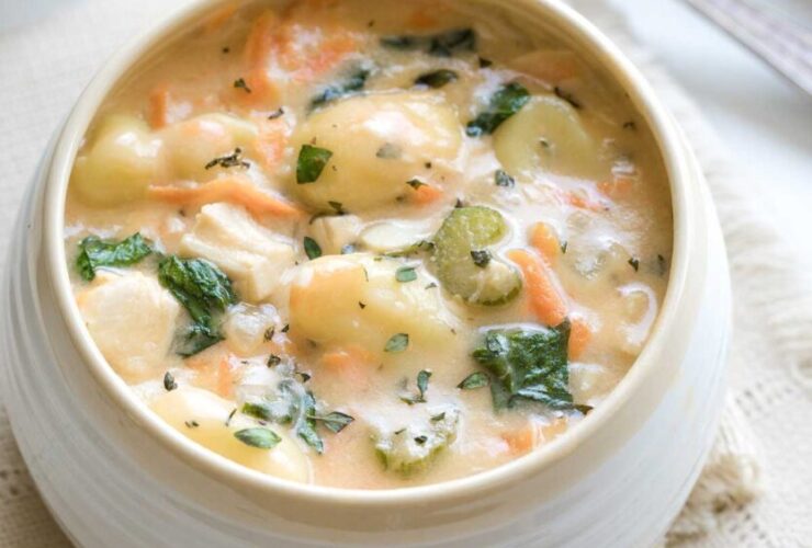 A bowl of creamy chicken gnocchi soup garnished with fresh thyme and a crack of black pepper, with soft potato gnocchi and shredded carrots visible in the rich cream broth, photographed on a rustic wooden table with a linen napkin