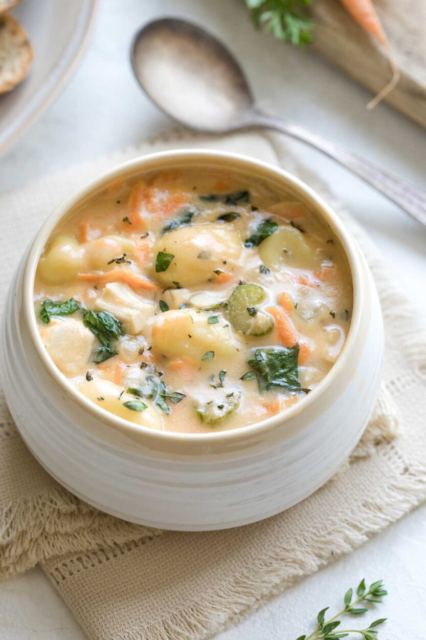 A bowl of creamy chicken gnocchi soup garnished with fresh thyme and a crack of black pepper, with soft potato gnocchi and shredded carrots visible in the rich cream broth, photographed on a rustic wooden table with a linen napkin
