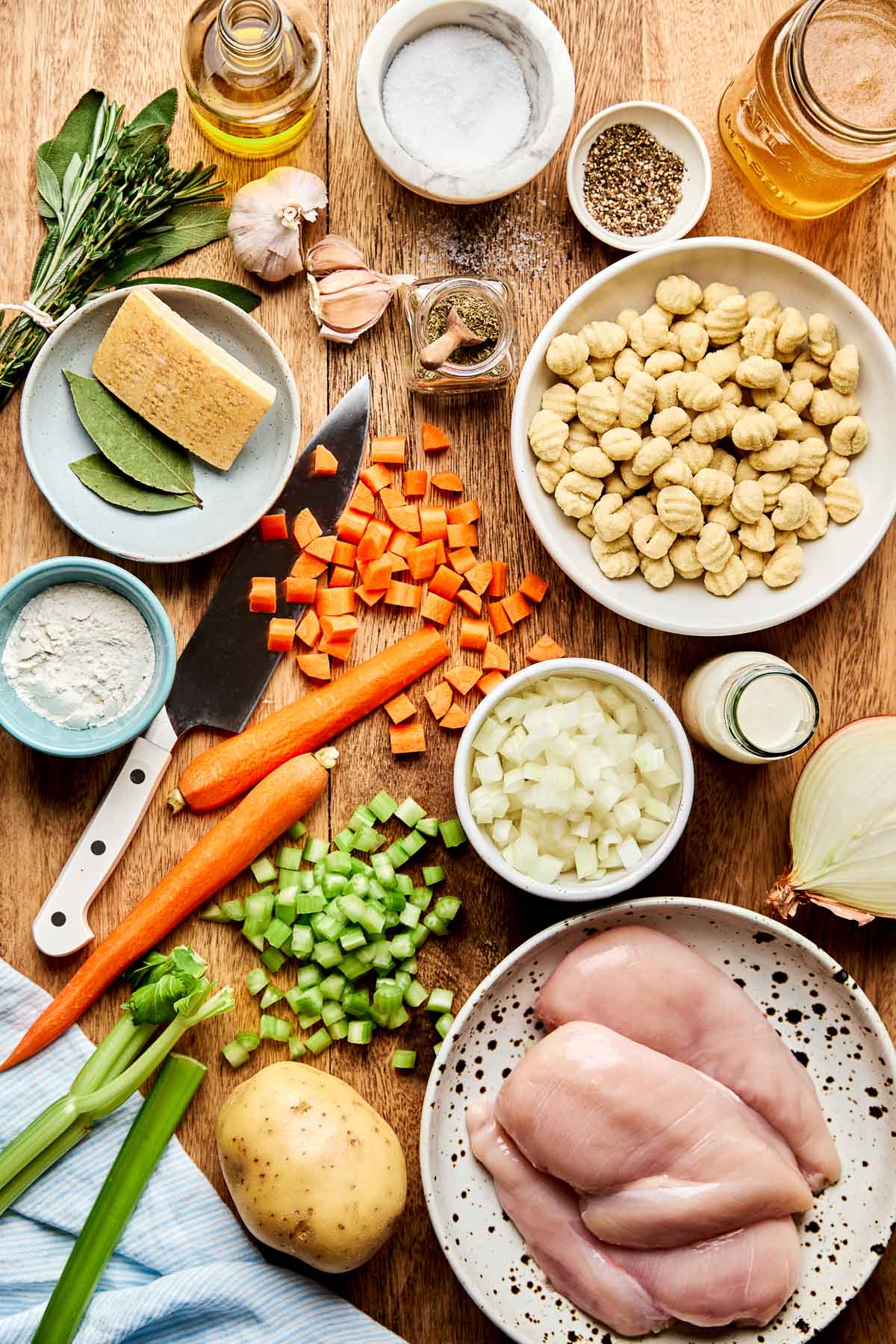 An overhead shot of the key ingredients for chicken gnocchi soup laid out on a marble surface: raw gnocchi, shredded rotisserie chicken, carrots, celery, garlic, onion, fresh thyme, heavy cream, and a carton of chicken broth