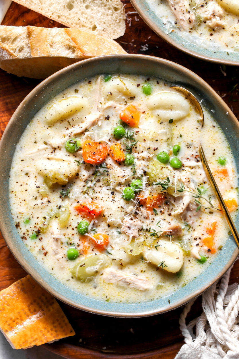 A close-up overhead shot of a rustic bowl of chicken gnocchi soup with a spoon resting in it, showing the pillowy gnocchi, shredded chicken, and vibrant shredded carrots in the thick cream broth, with crusty bread visible on the side