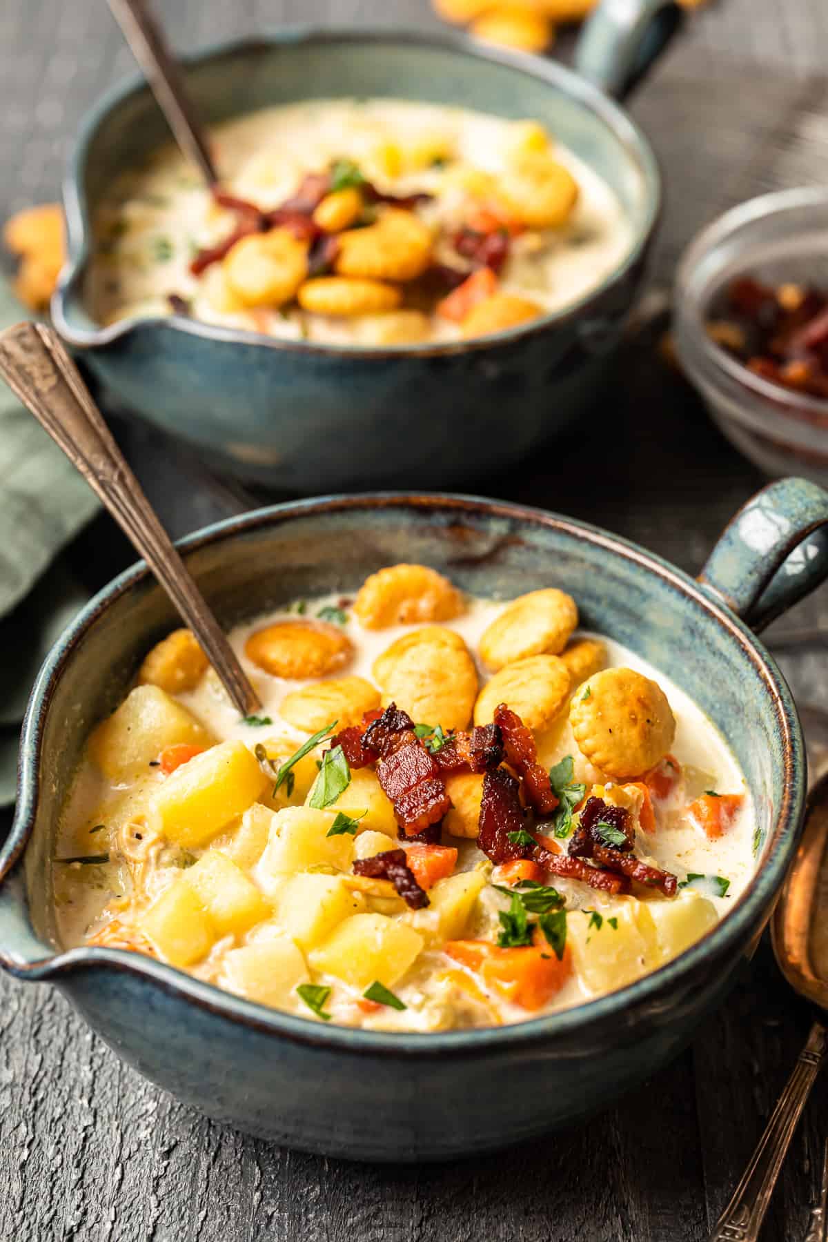 A rustic bowl of thick New England clam chowder topped with crispy bacon bits, fresh chives, and oyster crackers, served on a wooden table