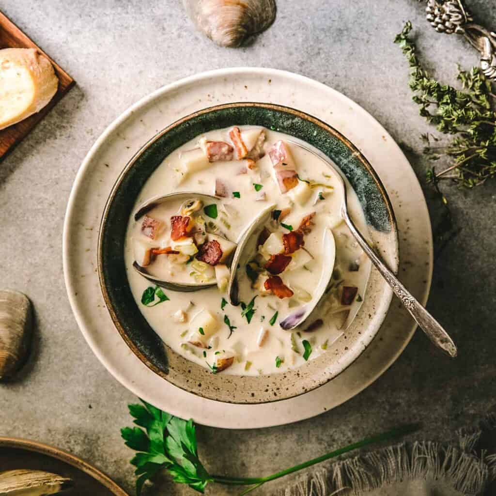 Close-up overhead shot of a bowl of clam chowder with crispy bacon, chives, oyster crackers, and a swirl of cream against a dark linen backdrop