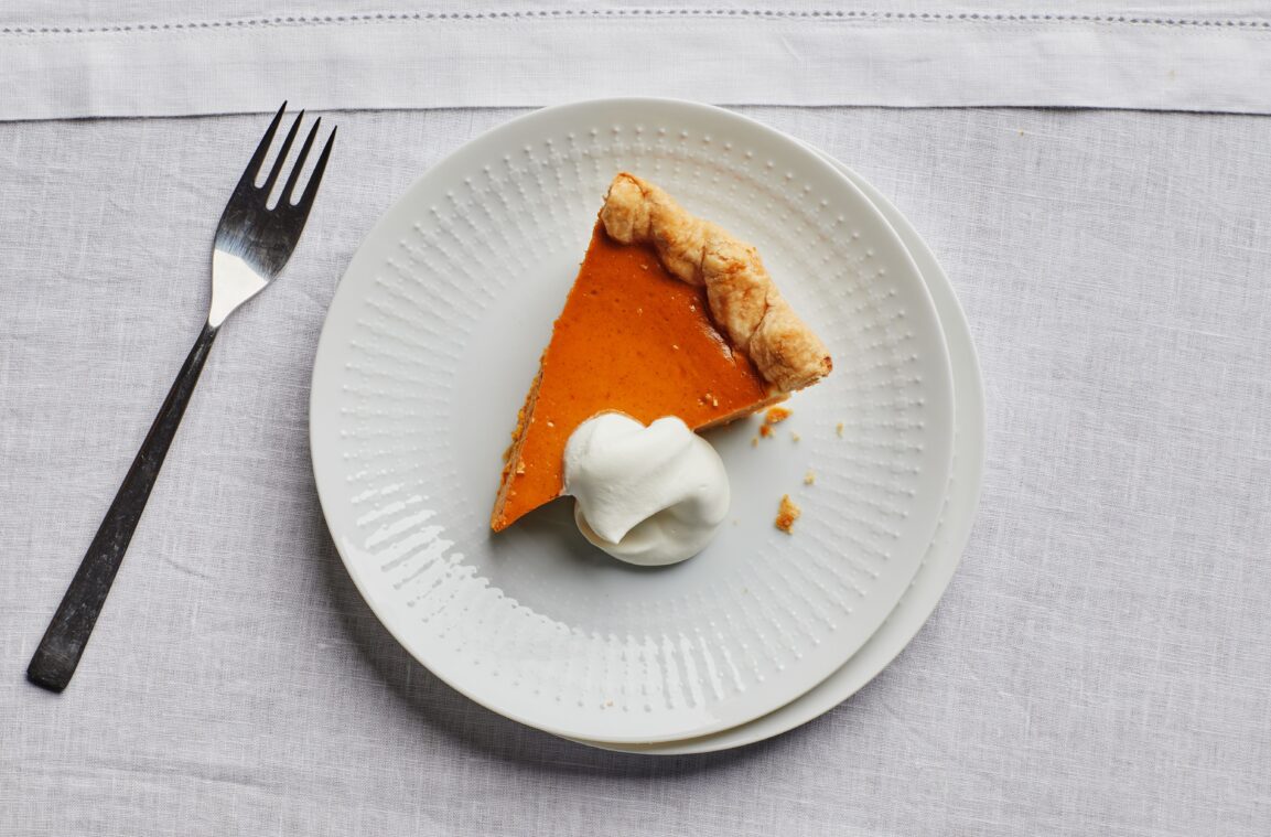 Close-up of a pumpkin pie slice on a fork, showing the smooth custard interior and flaky butter crust layers, whipped cream on top