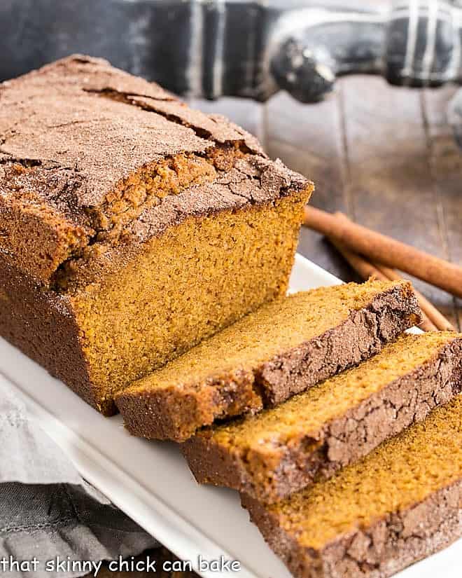 Sliced pumpkin bread with crackly cinnamon sugar top on a wooden cutting board, surrounded by fall spices and a linen napkin