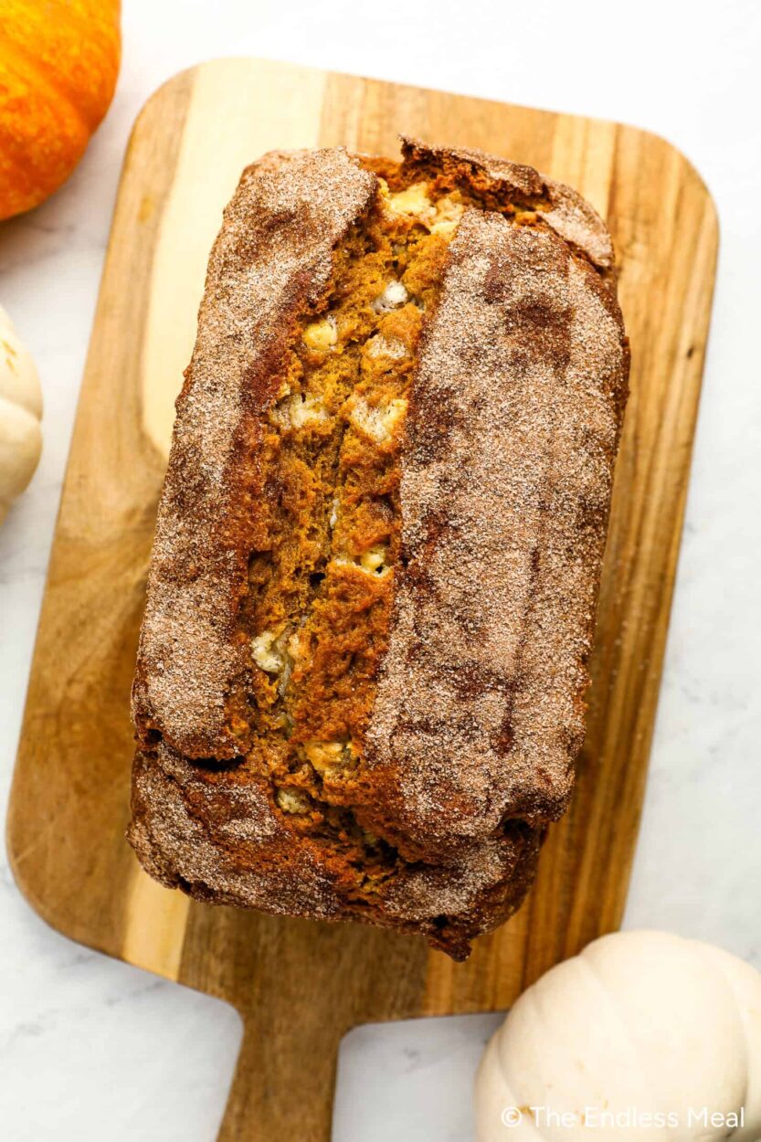 Overhead shot of pumpkin bread batter in a loaf pan with cinnamon sugar being spooned over the top, alongside measured spices and pumpkin purée