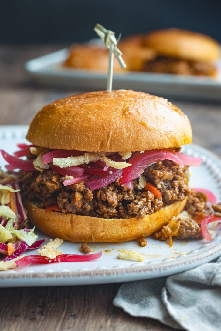 Classic homemade sloppy joes piled high on a toasted brioche bun, glossy sweet and tangy meat sauce dripping down the sides, served on a rustic wooden board with pickles and chips