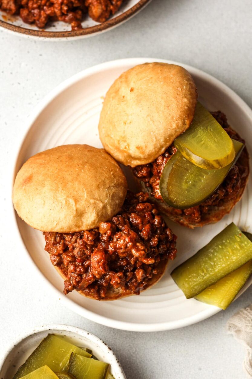 Close-up side-angle shot of a sloppy joe sandwich on a toasted brioche bun, glossy meat sauce spilling over the edges, garnished with a thin pickle slice, served with a pile of ridged potato chips on a dark plate