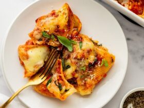 Overhead shot of jumbo pasta shells being filled with creamy ricotta mixture alongside small bowls of mozzarella, Parmesan, and fresh herbs