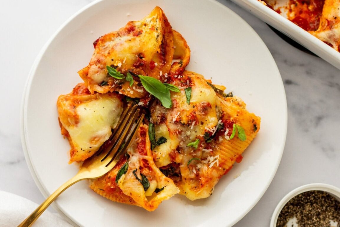 Overhead shot of jumbo pasta shells being filled with creamy ricotta mixture alongside small bowls of mozzarella, Parmesan, and fresh herbs
